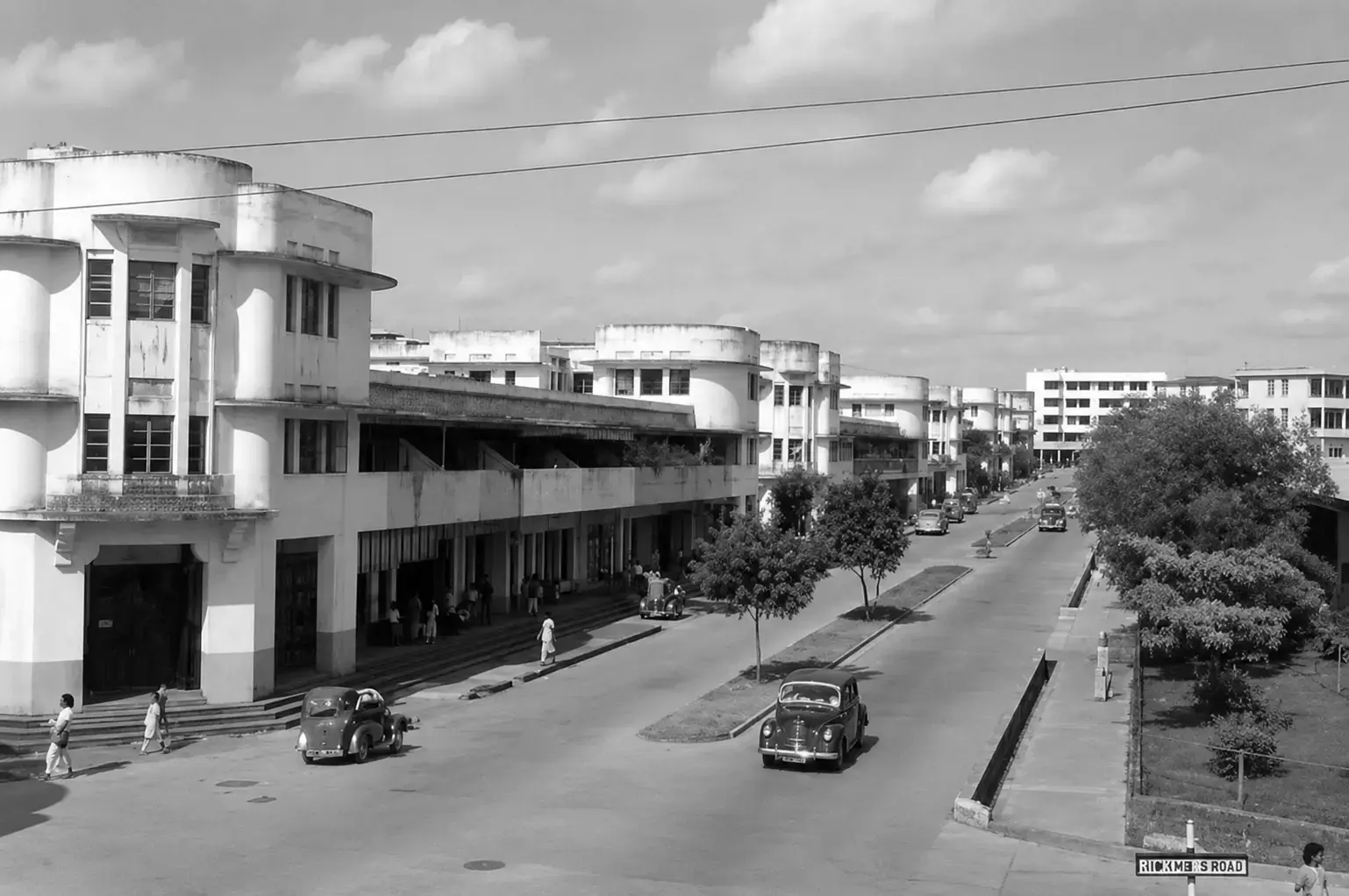A black-and-white historic photograph of a wide urban street, featuring Art Deco style buildings with rounded balconies lining the left side, vintage cars driving along the road, and a sign in the bottom right corner reading "RICKMERS ROAD".