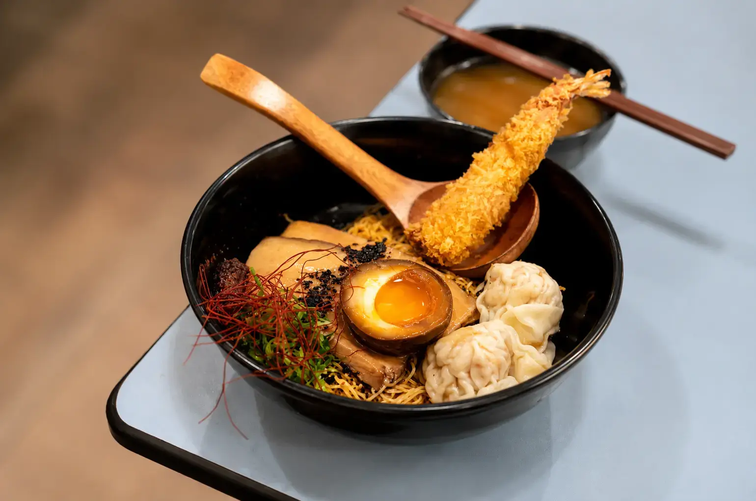 A bowl of dry noodles topped with a fried tempura prawn, soft-boiled egg, dumplings, and sliced pork belly, served alongside a small bowl of broth.