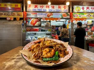 "A plate of Char Kway Teow with prawns and vegetables sits on a wooden table in front of a busy hawker center food stall labeled 'Lai Heng Fried Kuay Teow & Cooked Food'."