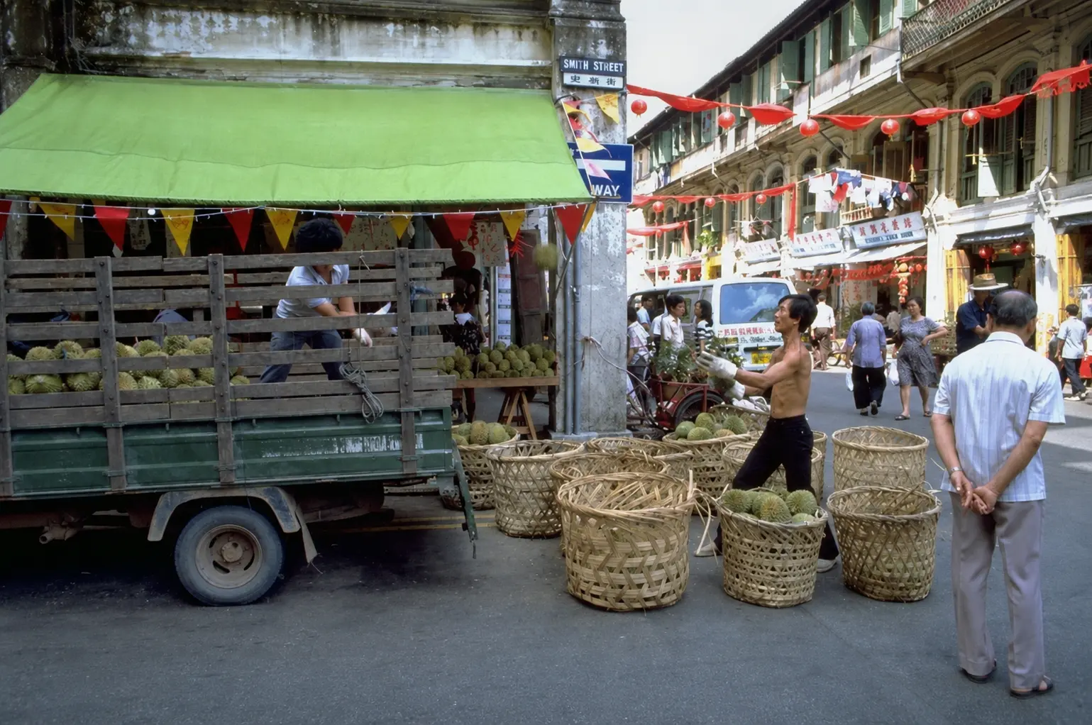 workers unload fresh durian fruit from a green truck on a Smith Street corner, tossing them into large woven baskets on the ground.