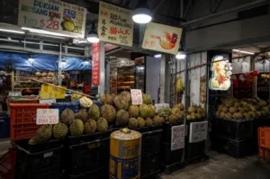 a vibrant street market stall at night displays large piles of whole, spiky durian fruit in plastic crates under bright hanging lights, with descriptive pricing signs for different varieties like Musang King, D24, and Red Prawn.