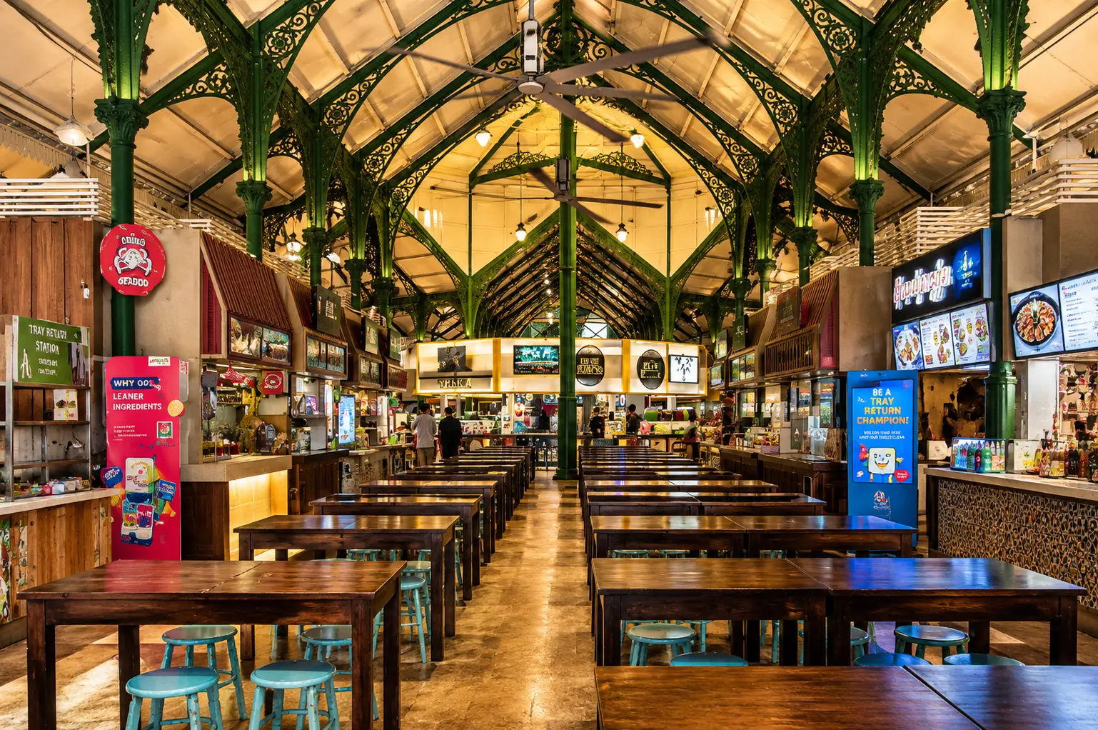 An interior view of Lau Pa Sat food hall in Singapore, featuring empty wooden dining tables, blue stools, ornate green iron arches, and surrounding food stalls.