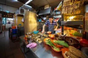A bustling indoor food court or hawker center (Lau Pa Sat) filled with people dining at round tables. The space features a high, vaulted ceiling supported by ornate green cast-iron columns and intricate archways. Food stalls line the sides under a warm, brightly lit atmosphere.