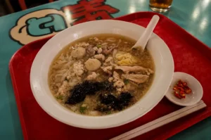 A large white bowl of noodle soup served on a red tray, topped with meatballs, minced meat, pork slices, and seaweed, with a side of sliced red chilies and chopsticks.