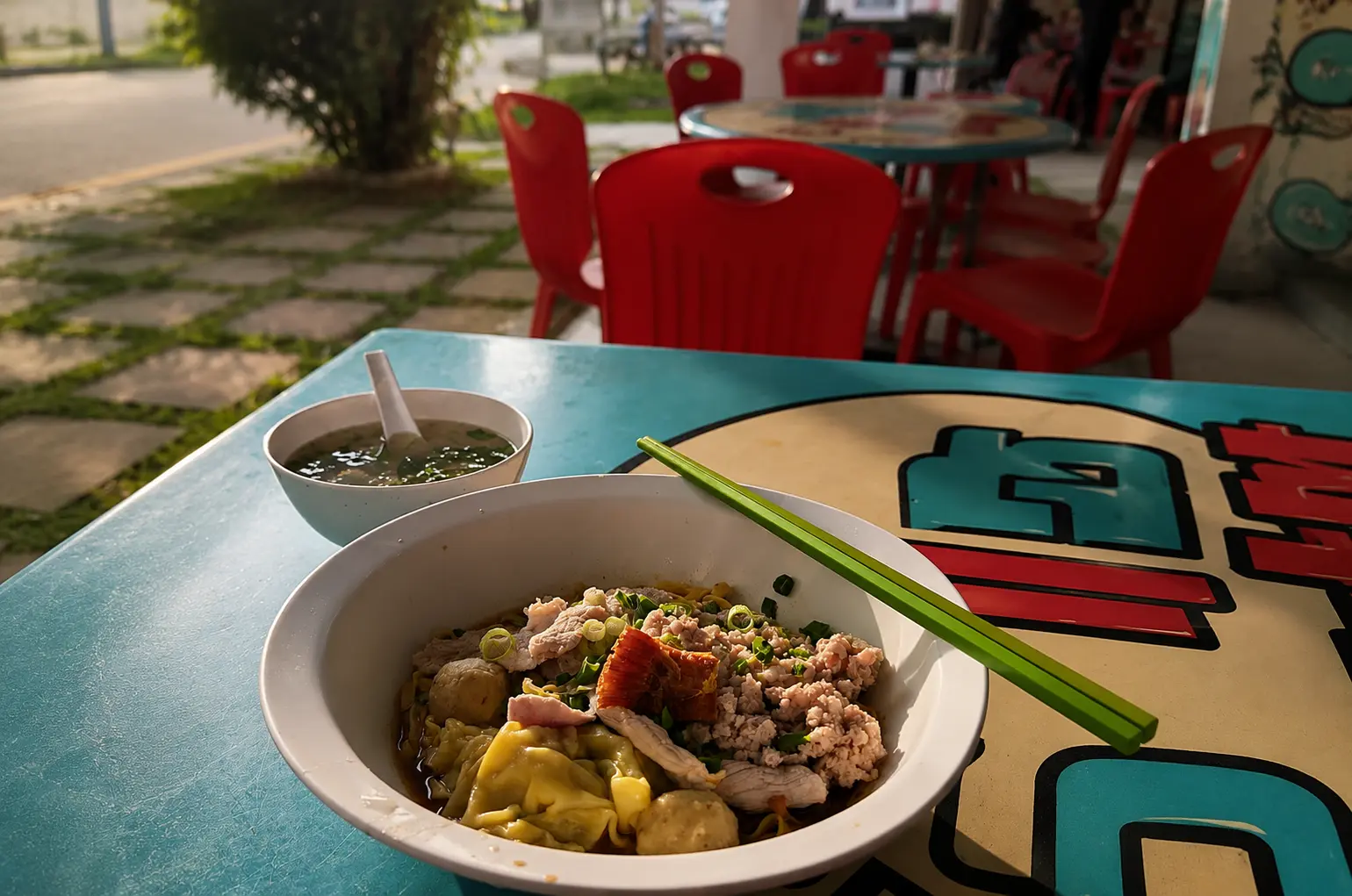 A bowl of dry noodles topped with minced pork, wontons, and meatballs, resting on a blue outdoor table with green chopsticks and a side bowl of broth, with blurry red chairs in the background.