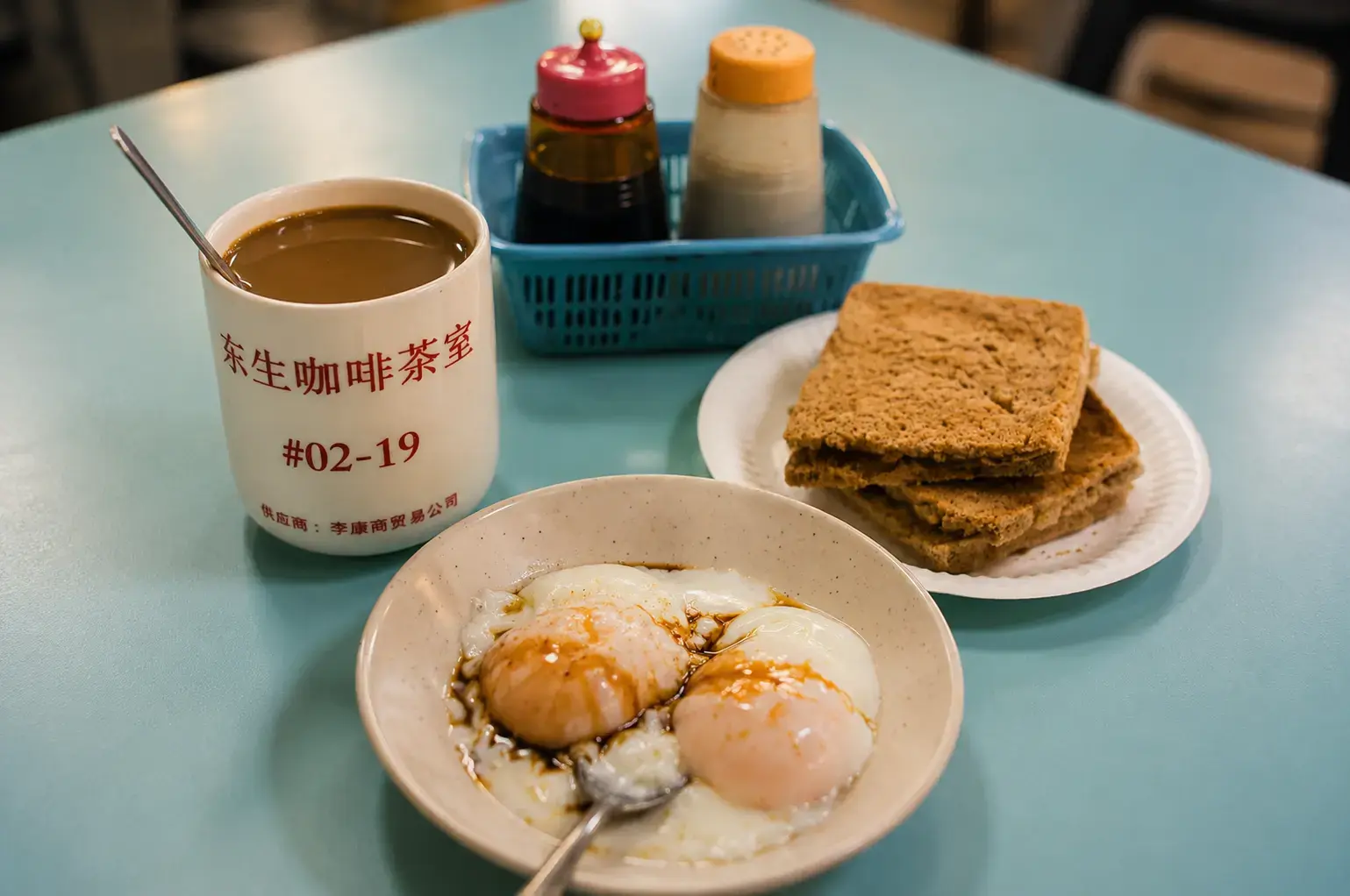 A traditional breakfast set on a light blue table featuring a mug of coffee, soft-boiled eggs with soy sauce, and toast.